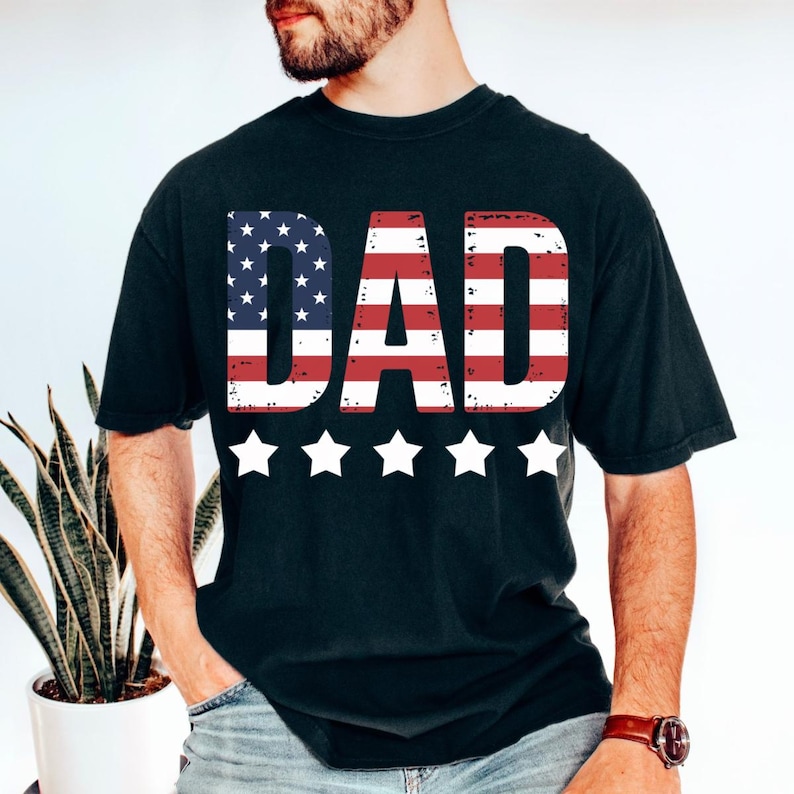 Puede incluir: Camiseta negra con la palabra "DAD" en rayas rojas y blancas y estrellas azules, que recuerda a la bandera americana. Cinco estrellas blancas est&aacute;n debajo. La camiseta es de estilo casual.