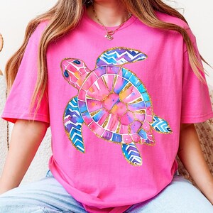 K&ouml;nnte beinhalten: Ein leuchtend pinkfarbenes T-Shirt mit einem farbenfrohen Aquarell-Design einer Meeresschildkr&ouml;te. Die Schildkr&ouml;te hat einen goldenen Umriss und ist mit Rosa-, Blau- und Gelbt&ouml;nen gef&uuml;llt. Das Shirt ist ein l&auml;ssiger Stil mit kurzen &Auml;rmeln.