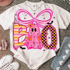 Op de afbeelding: Een wit t-shirt met een Halloween-thema. Het ontwerp spelt "BOO" met een roze spook, een strik en een geblokte "O". Het spook houdt vredestekens omhoog. Spinnen en spinnenwebben zijn ook te zien in het ontwerp.