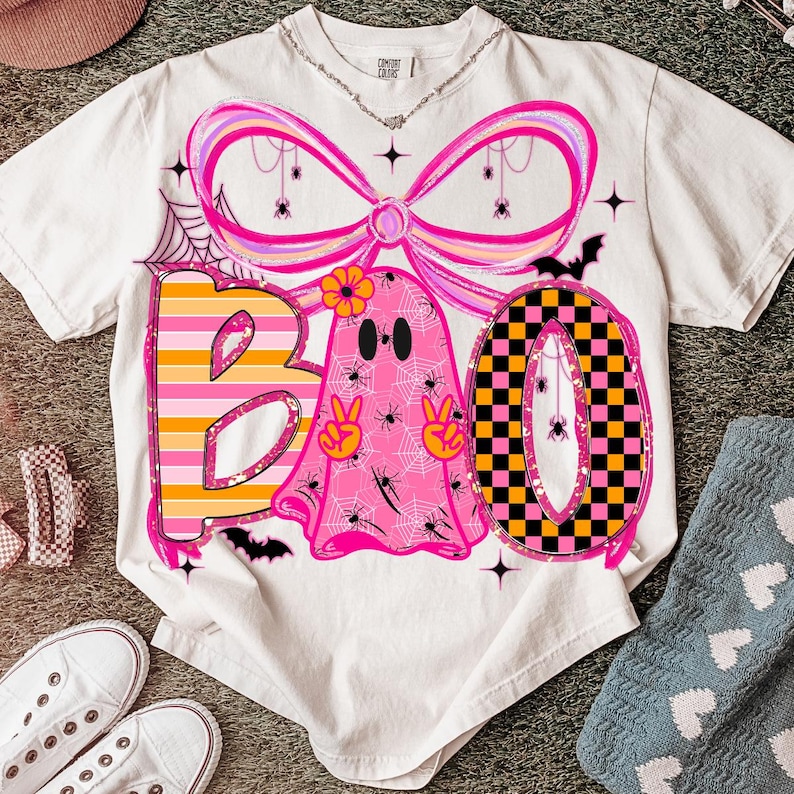 Op de afbeelding: Een wit t-shirt met een Halloween-thema. Het ontwerp spelt "BOO" met een roze spook, een strik en een geblokte "O". Het spook houdt vredestekens omhoog. Spinnen en spinnenwebben zijn ook te zien in het ontwerp.