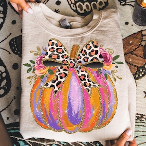 May include: A folded beige sweatshirt featuring a colorful pumpkin design. The pumpkin is decorated with orange, purple, and pink hues, accented with gold glitter. A leopard print bow and floral accents complete the festive design.