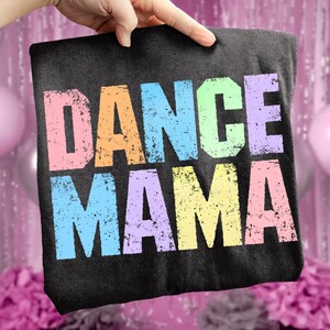 May include: A black t-shirt with the words "DANCE MAMA" printed in large, distressed, multi-colored letters. The letters are in shades of pink, orange, blue, green, and yellow. The shirt is held up by a hand.