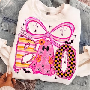 Op de afbeelding: Witte sweatshirt met een kleurrijk Halloween-thema. Het woord "BOO" wordt weergegeven met een gestreepte "B", een roze spookvormige "A" en een geblokte "O". Het ontwerp bevat spinnen, spinnenwebben en een grote roze strik.