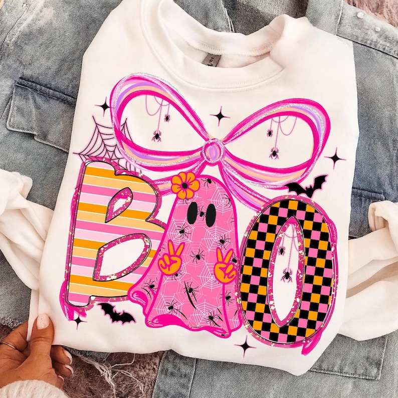 Op de afbeelding: Witte sweatshirt met een kleurrijk Halloween-thema. Het woord "BOO" wordt weergegeven met een gestreepte "B", een roze spookvormige "A" en een geblokte "O". Het ontwerp bevat spinnen, spinnenwebben en een grote roze strik.