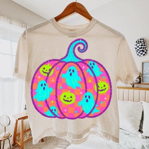 May include: Beige t-shirt with a Halloween-themed graphic. The design features a large pumpkin shape with a teal stem and a purple outline. Inside the pumpkin are teal ghosts and green jack-o'-lanterns. The pumpkin has a pink and orange striped pattern.