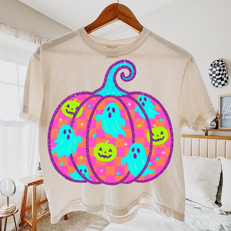 May include: Beige t-shirt with a Halloween-themed graphic. The design features a large pumpkin shape with a teal stem and a purple outline. Inside the pumpkin are teal ghosts and green jack-o'-lanterns. The pumpkin has a pink and orange striped pattern.