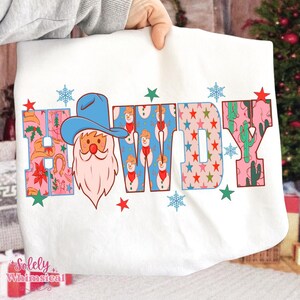K&ouml;nnte beinhalten: Wei&szlig;es Sweatshirt mit dem Wort "HOWDY" in bunten Buchstaben. Das "H" zeigt einen Weihnachtsmann mit Cowboyhut. Das "W" hat ein Schneemannmuster und das "Y" ein Kaktusdesign. Rote Sterne und blaue Schneeflocken sind um das Wort herum verstreut.