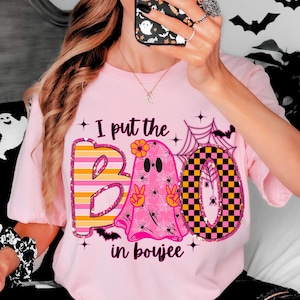 May include: Pink t-shirt with the text "I put the BOO in boujee." The word "BOO" is designed with a striped orange and yellow "B", a pink ghost with peace signs for hands, and a checkered black and yellow "O".
