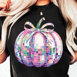 Puede incluir: Camiseta negra con un dise&ntilde;o de calabaza de bola de discoteca colorida. La calabaza est&aacute; hecha de peque&ntilde;os cuadrados reflectantes en tonos de rosa, morado, azul y verde. Un tallo verde y un lazo arco&iacute;ris completan el dise&ntilde;o festivo.