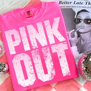 Puede incluir: Camiseta rosa brillante con las palabras "PINK OUT" en letras blancas grandes y desgastadas. La camiseta est&aacute; sobre una superficie a cuadros rosa y blanco, con una bola de discoteca y una revista en el fondo. La camiseta est&aacute; etiquetada como "Comfort Colors".