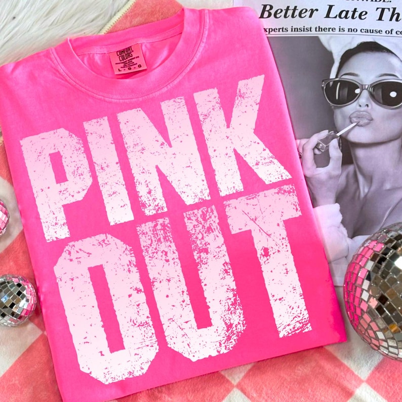Puede incluir: Camiseta rosa brillante con las palabras "PINK OUT" en letras blancas grandes y desgastadas. La camiseta est&aacute; sobre una superficie a cuadros rosa y blanco, con una bola de discoteca y una revista en el fondo. La camiseta est&aacute; etiquetada como "Comfort Colors".