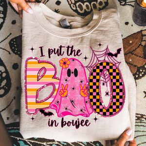 May include: A beige sweatshirt with the phrase "I put the BOO in boujee" in a colorful Halloween design. The "B" is striped, the "O" is a pink ghost, and the second "O" is checkered. Includes bats, spiders, and spiderwebs.