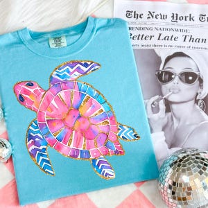 K&ouml;nnte beinhalten: Hellblaues T-Shirt mit einem farbenfrohen Aquarell-Meeresschildkr&ouml;ten-Design. Die Schildkr&ouml;te hat rosa, blaue und goldene Akzente. Das T-Shirt liegt neben einer Zeitung mit der &Uuml;berschrift "Better Late Than".