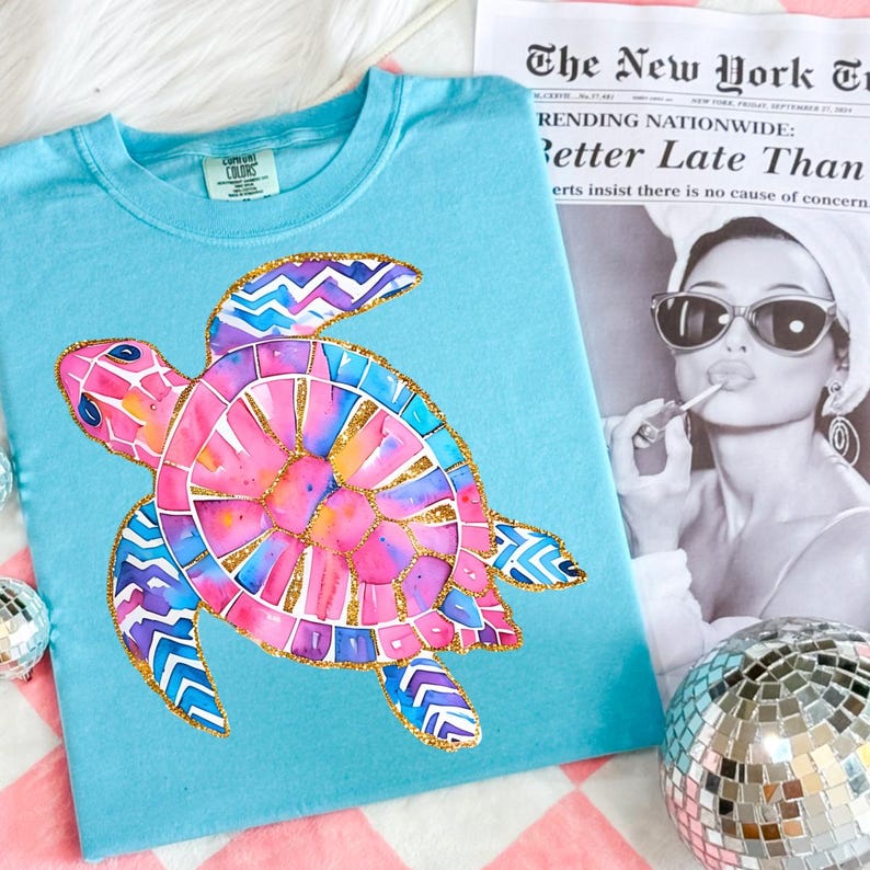 K&ouml;nnte beinhalten: Hellblaues T-Shirt mit einem farbenfrohen Aquarell-Meeresschildkr&ouml;ten-Design. Die Schildkr&ouml;te hat rosa, blaue und goldene Akzente. Das T-Shirt liegt neben einer Zeitung mit der &Uuml;berschrift "Better Late Than".