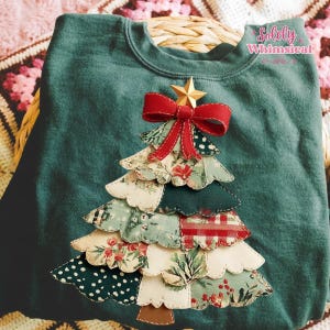 Puede incluir: Una sudadera verde azulado con un diseño de árbol de Navidad de tela. El árbol está hecho de varias telas estampadas, coronado con un lazo rojo y una estrella dorada. El texto "Solely Whimsical" es visible en la esquina superior derecha.