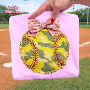 Pode incluir: Uma camiseta rosa claro com um design de softball. O softball é feito de tecido camuflado amarelo e verde com costuras vermelhas. Uma fita combinando está no topo. O texto "Solely Whimsical" está no canto superior esquerdo.