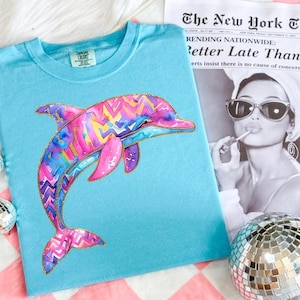 May include: A light blue t-shirt featuring a colorful watercolor dolphin design with pink, purple, and yellow accents. The dolphin has a gold glitter outline. The shirt is next to a black and white newspaper with the headline "Better Late Than".