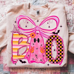 Op de afbeelding: Een beige sweatshirt met een kleurrijk Halloween-ontwerp. Het ontwerp bevat het woord "BOO" met een gestreepte "B", een roze spook en een geblokte "O". Het ontwerp bevat ook spinnen, vleermuizen en een grote roze strik.