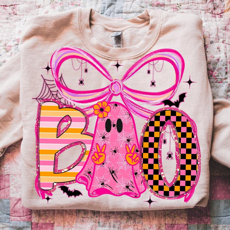 Op de afbeelding: Een beige sweatshirt met een kleurrijk Halloween-ontwerp. Het ontwerp bevat het woord "BOO" met een gestreepte "B", een roze spook en een geblokte "O". Het ontwerp bevat ook spinnen, vleermuizen en een grote roze strik.