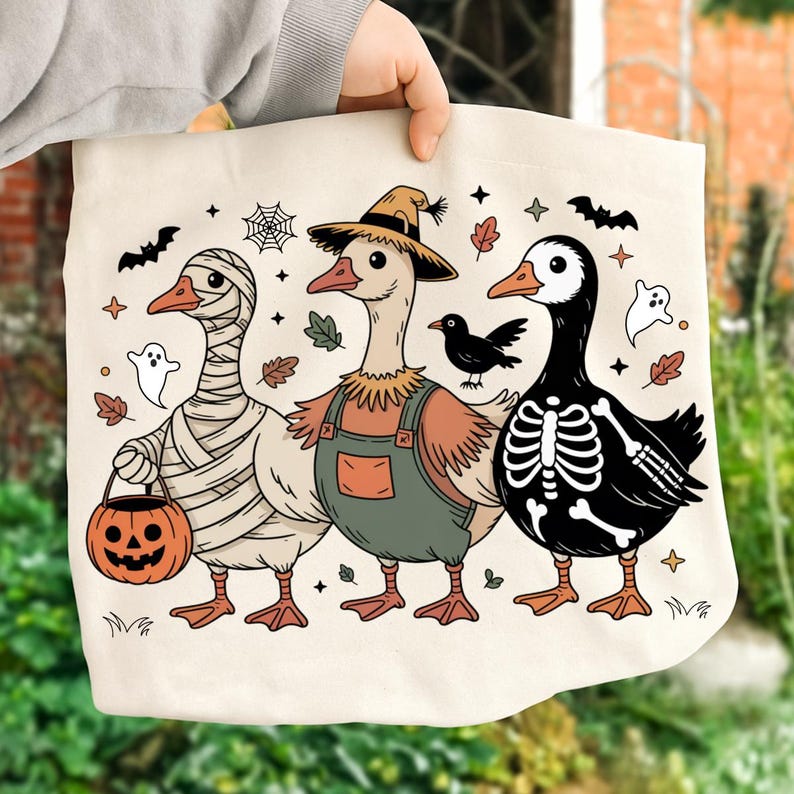 May include: A cream-colored tote bag featuring three geese in Halloween costumes. One is wrapped in bandages, another wears overalls and a straw hat, and the third is a skeleton. Bats, ghosts, and leaves decorate the background.