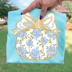 May include: A light blue t-shirt featuring a white pumpkin design with blue roses and green leaves. The pumpkin has a gold glitter outline and a matching bow. The text "Solely Whimsical" is visible in the upper right corner.