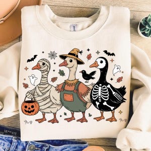 May include: Cream-colored sweatshirt featuring a Halloween-themed graphic of three geese in costumes. One is wrapped in bandages, another wears overalls and a straw hat, and the third is a skeleton. Includes bats, leaves, and a jack-o'-lantern.