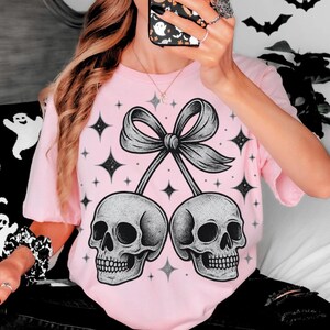 K&ouml;nnte beinhalten: Hellrosa T-Shirt mit einem Motiv aus zwei sch&auml;delartigen Kirschen, die durch eine Schleife verbunden sind. Das Design beinhaltet Sterne. Das T-Shirt hat eine hellrosa Farbe.