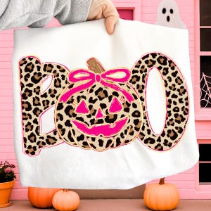 Op de afbeelding: Wit Halloween-thema handdoek met het woord "BOO" in luipaardprint. Een roze pompoen met een strik staat in het midden. De handdoek wordt door een persoon omhoog gehouden. Oranje pompoenen staan onderaan.