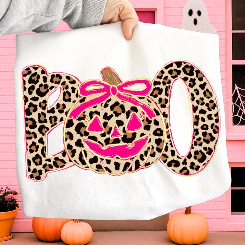 Op de afbeelding: Wit Halloween-thema handdoek met het woord "BOO" in luipaardprint. Een roze pompoen met een strik staat in het midden. De handdoek wordt door een persoon omhoog gehouden. Oranje pompoenen staan onderaan.