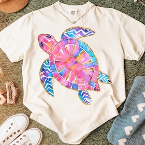 K&ouml;nnte beinhalten: Cremefarbenes T-Shirt mit einem farbenfrohen Aquarell-Schildkr&ouml;ten-Design. Die Schildkr&ouml;te hat rosa, blaue und lila Farbt&ouml;ne mit goldenen Akzenten. Eine zarte Silberkette ist sichtbar. Teil einer Casual-Wear-Kollektion.