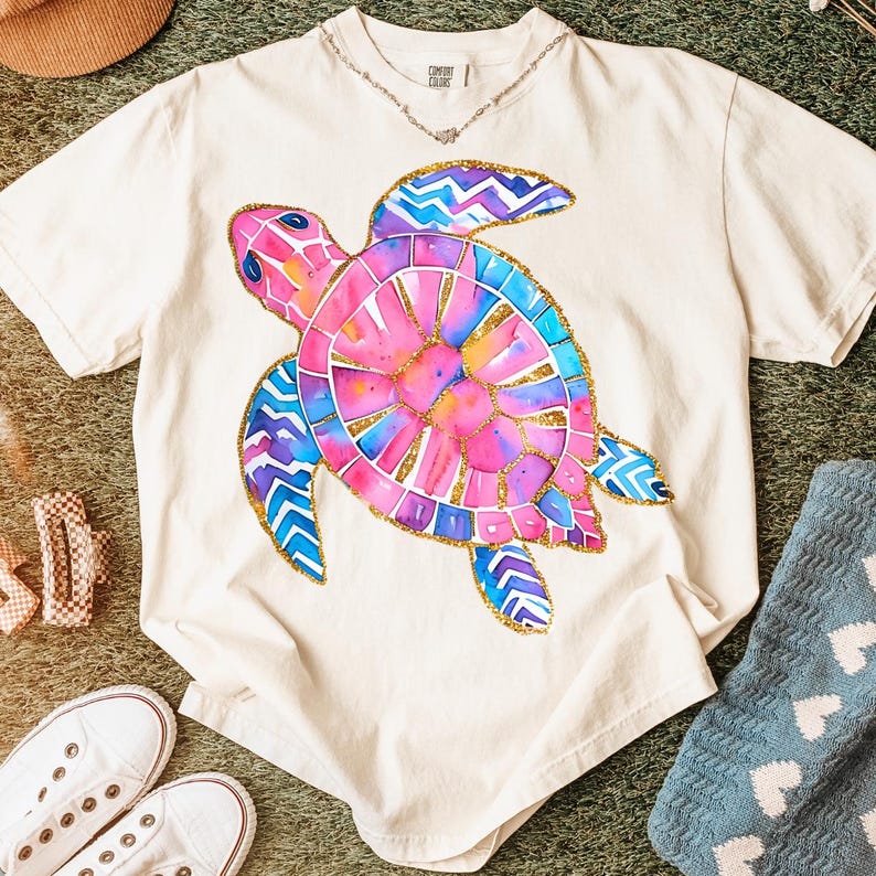 K&ouml;nnte beinhalten: Cremefarbenes T-Shirt mit einem farbenfrohen Aquarell-Schildkr&ouml;ten-Design. Die Schildkr&ouml;te hat rosa, blaue und lila Farbt&ouml;ne mit goldenen Akzenten. Eine zarte Silberkette ist sichtbar. Teil einer Casual-Wear-Kollektion.
