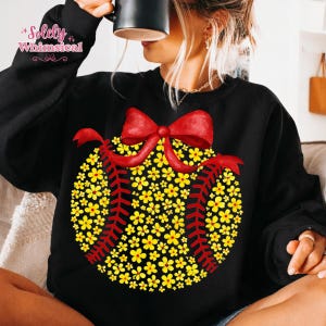 May include: Black sweatshirt featuring a baseball design made of yellow flowers and red stitching. A large red bow is at the top of the baseball. The text "Solely Whimsical" is in the upper left corner.