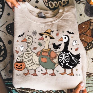 May include: Beige sweatshirt featuring three cartoon geese in Halloween costumes. One is wrapped in bandages, another wears overalls and a scarecrow hat, and the third is a skeleton. Includes bats, ghosts, and a jack-o'-lantern.