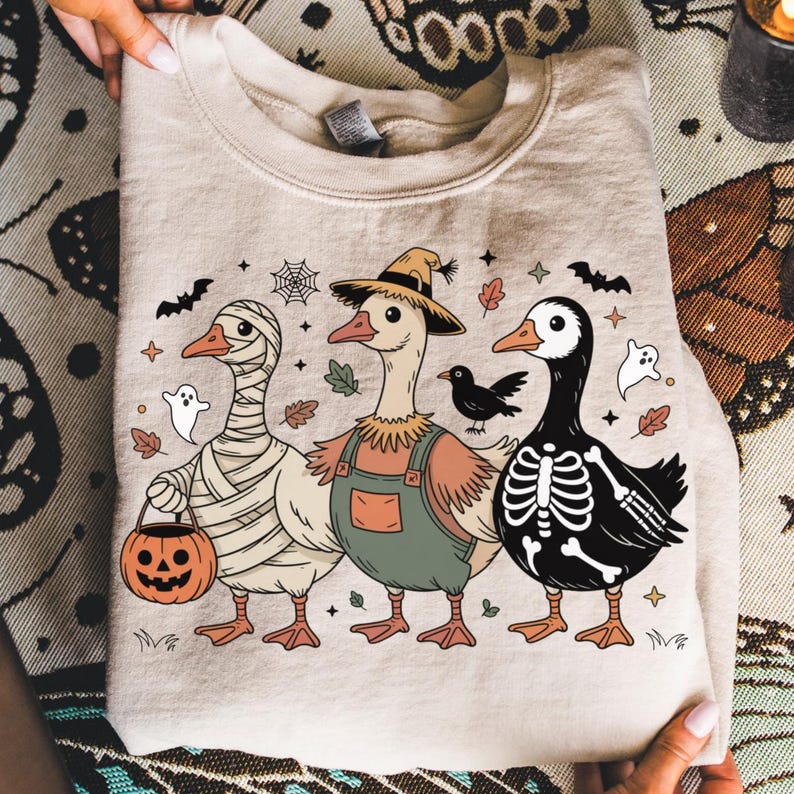 May include: Beige sweatshirt featuring three cartoon geese in Halloween costumes. One is wrapped in bandages, another wears overalls and a scarecrow hat, and the third is a skeleton. Includes bats, ghosts, and a jack-o'-lantern.