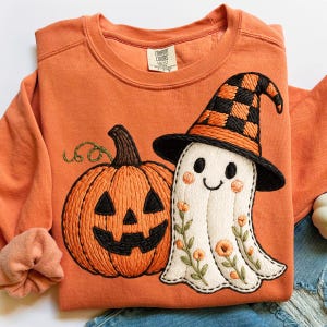 May include: Orange sweatshirt with embroidered Halloween design. Features a jack-o'-lantern and a ghost wearing a witch hat. The ghost is decorated with floral accents. The sweatshirt is a Comfort Colors brand.