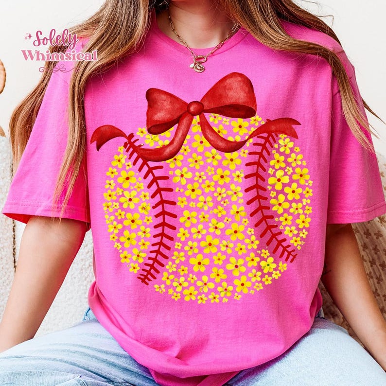May include: A pink t-shirt featuring a baseball design. The baseball is filled with yellow flowers and red stitching. A red bow is at the top. The text "Solely Whimsical" is in the upper left corner.