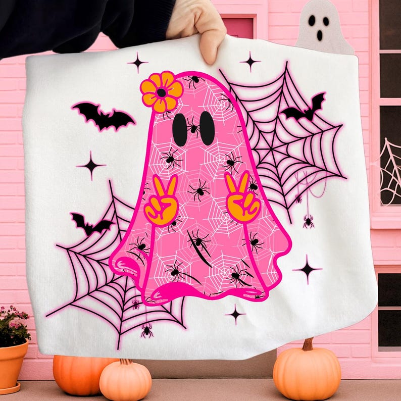 Pode incluir: Um cobertor branco com tema de Halloween com um fantasma rosa com uma flor e sinais de paz. O fantasma &eacute; decorado com aranhas e teias de aranha. Morcegos e estrelas pretas tamb&eacute;m est&atilde;o presentes no design. O cobertor &eacute; segurado em frente a um fundo rosa.
