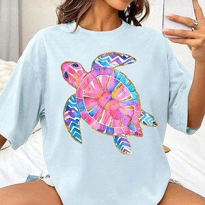 K&ouml;nnte beinhalten: Hellblaues T-Shirt mit einem farbenfrohen Aquarell-Meeresschildkr&ouml;ten-Design. Die Schildkr&ouml;te hat rosa, blaue und lila Farbt&ouml;ne mit goldenen Akzenten. Das Shirt hat eine l&auml;ssige, &uuml;bergro&szlig;e Passform.