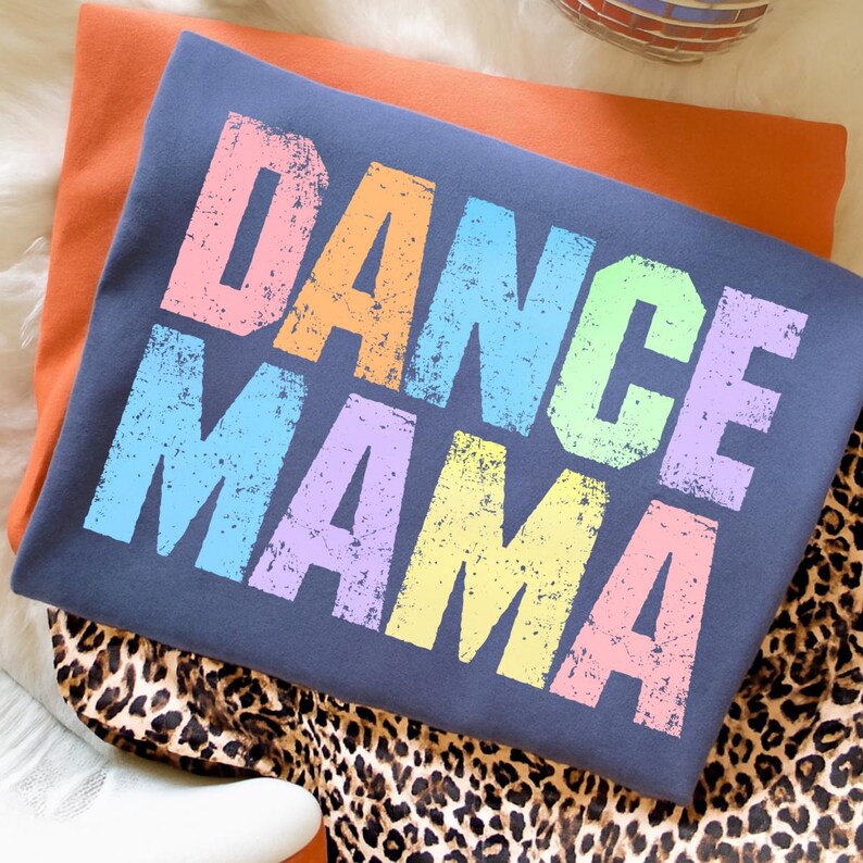 May include: A dark blue t-shirt with the words "DANCE MAMA" in large, colorful, distressed letters. The shirt is folded on top of an orange shirt and a leopard print fabric.
