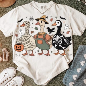 May include: Cream-colored t-shirt featuring three cartoon geese in Halloween costumes. One is wrapped in bandages, another wears overalls and a straw hat, and the third is a skeleton. The shirt has a Comfort Colors tag.