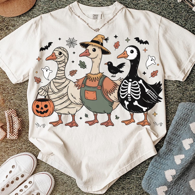 May include: Cream-colored t-shirt featuring three cartoon geese in Halloween costumes. One is wrapped in bandages, another wears overalls and a straw hat, and the third is a skeleton. The shirt has a Comfort Colors tag.