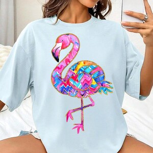 May include: Light blue t-shirt featuring a colorful flamingo design. The flamingo has a pink head and neck, a black beak, and a body with a vibrant, patterned design in shades of pink, blue, yellow, and purple. The design is outlined with gold glitter.