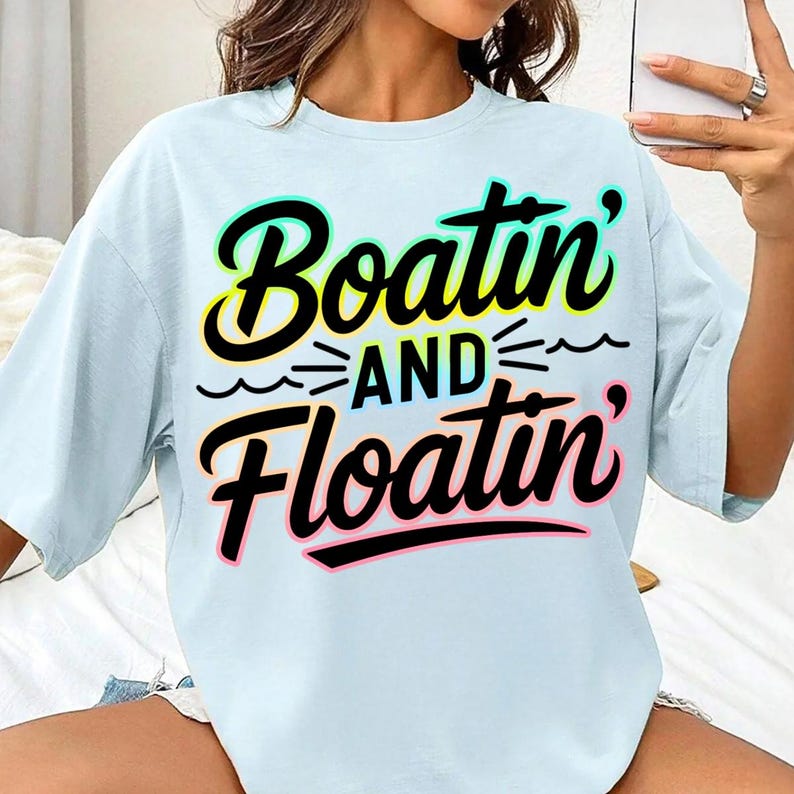K&ouml;nnte beinhalten: Hellblaues T-Shirt mit dem Schriftzug "Boatin' and Floatin'" in einer farbenfrohen, verspielten Schriftart. Der Text ist schwarz umrandet und mit gelben, gr&uuml;nen und rosa Akzenten versehen. Das Shirt hat eine lockere Passform und kurze &Auml;rmel.