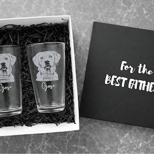 May include: A gift set with two clear pint glasses, each etched with a Labrador Retriever portrait and the name "Goose." The glasses are in a white box with black filler, next to a black box lid with the text "For the BEST FATHER."