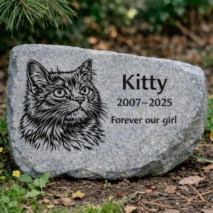 May include: A gray stone pet memorial with a detailed black and white illustration of a cat's face. The name "Kitty" is inscribed, along with the years 2007-2025 and the phrase "Forever our girl."