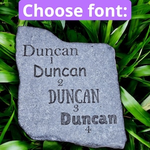 Op de afbeelding: Een grijze steen met de naam "Duncan" vier keer in verschillende lettertypen gegraveerd. De tekst "Choose font:" staat in een paarse rechthoek bovenaan. De steen staat tegen een achtergrond van groen gebladerte.