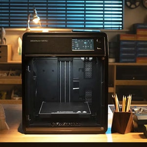 May include: A black 3D printer with a glass door, showing the interior with a build plate. The printer is labeled "Adventurer 3 Lite Pro" and is sitting on a wooden table with other 3D printed objects.