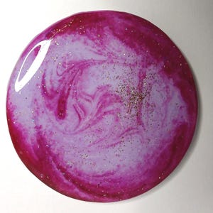 May include: A round, decorative coaster with a swirling design in shades of pink and lavender. The coaster has a glossy finish and is speckled with glitter for added sparkle. The design creates a marbled effect.