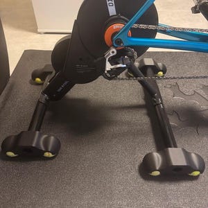 Op de afbeelding: Een zwarte en blauwe fietstrainer met een zwart vliegwiel en ketting. De trainer heeft een zwarte basis met gele accenten en de tekst "D100" en "ZWIFT" zijn zichtbaar.