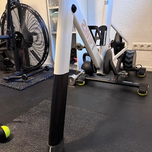 Puede incluir: Bicicleta estática blanca y negra con la palabra "ZWIFT" impresa en el marco. Cuenta con un portabotellas y un ventilador negro en el fondo. La bicicleta está sobre una esterilla negra.